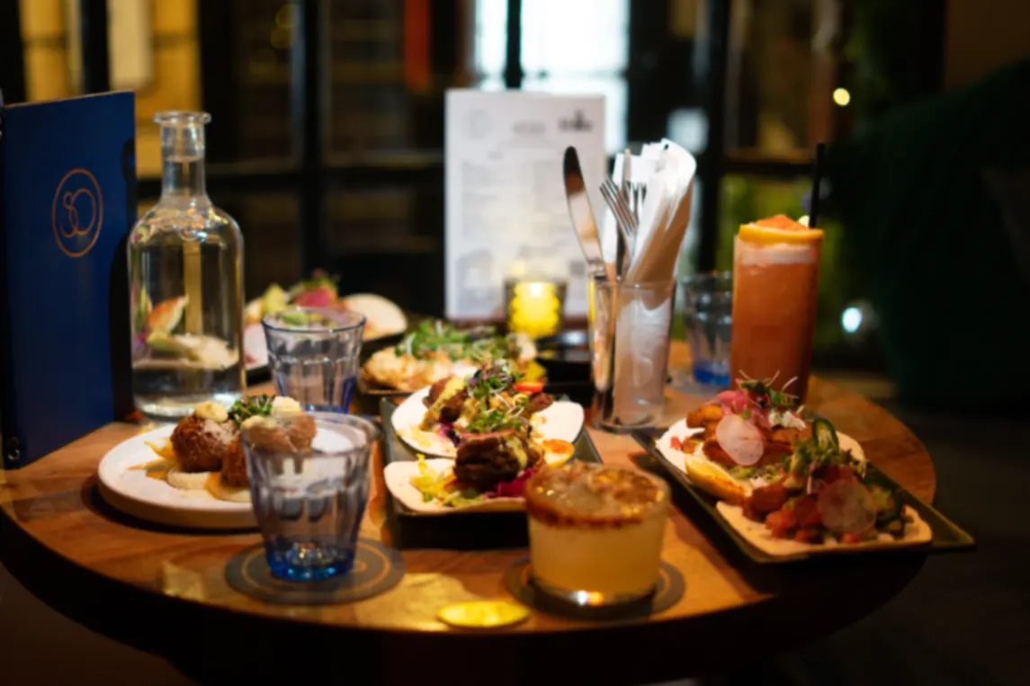 Round table set with sharing plates, cocktails and menu in a dim-lit restaurant